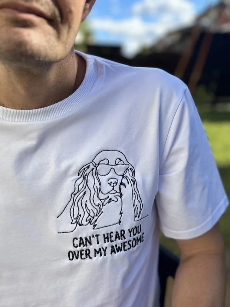 A man in a white t-shirt embroidered with the spaniel design and the words CAN’T HEAR YOU OVER MY AWESOME